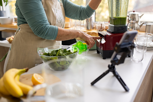 Senior woman filming herself with smart phone while she is preparing a healthy meal in a blender in kitchen at home.