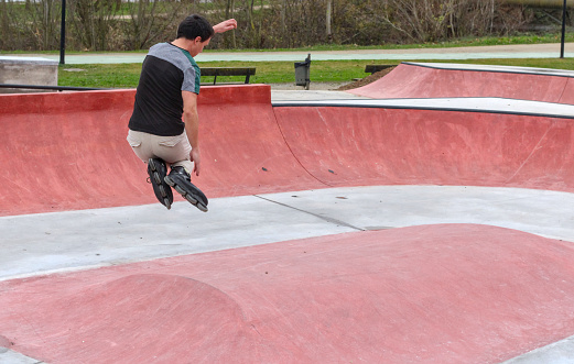 Young white guy with black roller skates in a skate park, dark hair, no helmet, making a jump in the air, with dynamic movement, natural background. Copy space