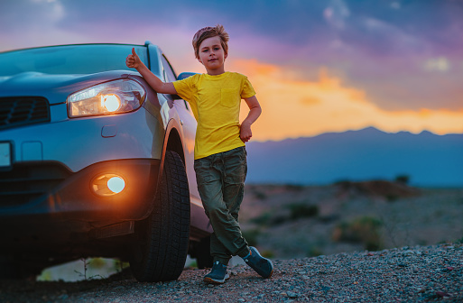 Successful boy standing by the car and showing thumbs up at twilight on mountains background