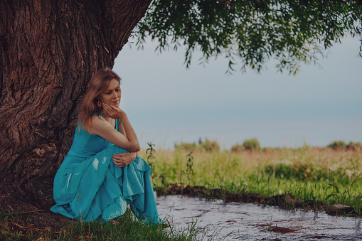 Thoughtful elegant woman in blue dress sitting under tree by a stream