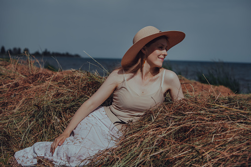 Beautiful young woman in hat resting on haystack on the shore of lake