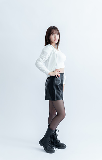 Young Asian woman in a white crop top, black leather skirt, and black boots posing on a white background