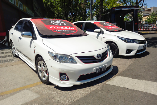 Buenos Aires, Argentina - Nov 6, 2022: Two compact Toyota Corolla 2000s y 2010s sedan in the street at a classic car show. Flag with badge and branding covering the windscreen.