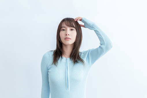 A young Asian woman in a blue sweater is standing with her hand on her head and looking at the camera with a serious expression.