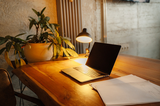 Front view of a wooden desk with a laptop and a plant at the left of the image and and a desk lamp at the center. The image is illuminated by the desk lamp.