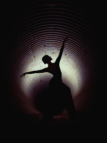 Silhouette of faceless woman dancing with raised arms in dark underground tunnel illuminated by light