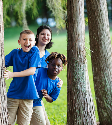 Two multiracial boys having fun at summer camp, standing between tree trunks in the park with their camp counselor, a young Hispanic and Native American woman. The main focus is on the 11 year old African-American boy. They are looking toward the camera.