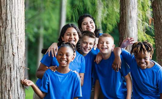 A multiracial group of five children and their camp counselor having fun at summer camp. They are standing together by trees at the park.