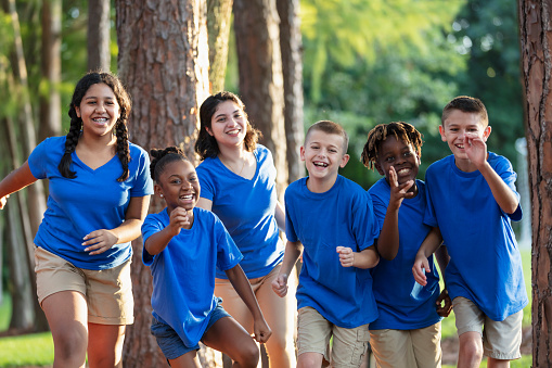 A multiracial group of five children and their camp counselor having fun at summer camp. The children, 10 to 12 years old, are at the park surrounded by trees, running toward the camera.
