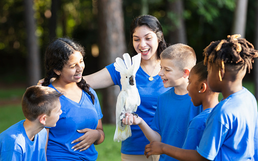 A multiracial group of five children and their camp counselor on a field trip to a park during summer camp. One of the boys is holding a cockatoo.