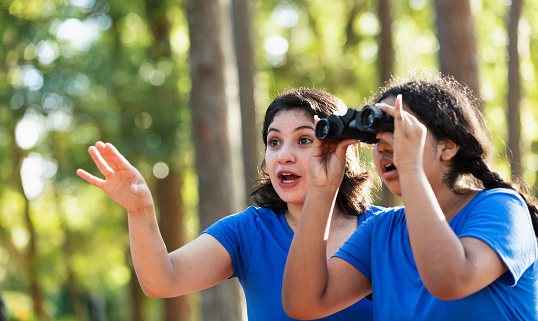 An Hispanic girl on a field trip in a park, with a pair of binoculars. She and a teacher or summer camp counselor look amazed by what they see.