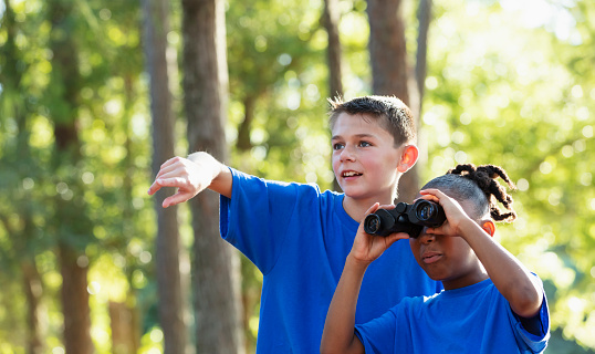 Two multiracial children on a field trip to the park on a sunny day, exploring with a pair of binoculars.