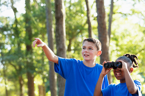 Two multiracial children on a field trip to the park on a sunny day, exploring with a pair of binoculars.