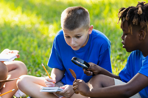 Two multiracial boys learning about the environment in their outdoor classroom. A 10 year old boy is sitting cross-legged on the grass taking notes. His African-American friend is examining a leaf through a magnifying glass.