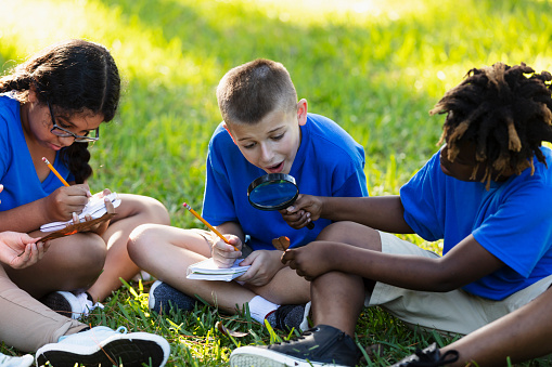 A group of three multiracial 10 year old children learning about the environment in their outdoor classroom. One of the boys is amazed, looking at a leaf through a magnifying glass his African-American friend is holding. An Hispanic girl is writing on a note pad.