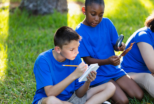Two multiracial children learning about the environment in their outdoor classroom. A 12 year old boy is sitting cross-legged on the grass taking notes. His friend, a 10 year old African-American girl, examines a leaf through a magnifying glass.