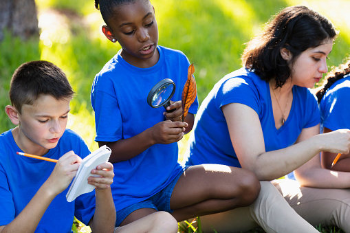 Multiracial children learning about the environment in their outdoor classroom. A 12 year old boy is sitting cross-legged on the grass taking notes. His friend, a 10 year old African-American girl, examines a leaf through a magnifying glass.