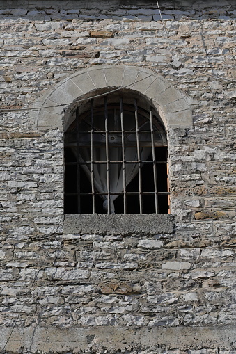 Round arch and semicircular transom barred window with cast-iron grilles and folded white curtain cut into an Ottoman-era house stone wall in the Clock Tower area of the citadel. Gjirokaster-Albania.