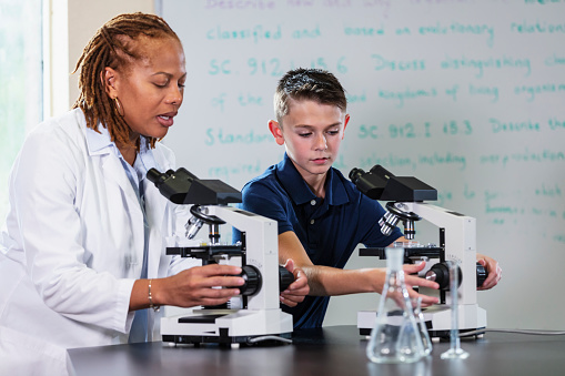 A science teacher in elementary or middle school showing a student how to use a microscope. The teacher is a mature multiracial woman, in her 40s. Her student is a 12 year old boy.
