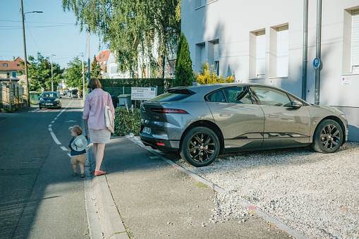 Paris, France - Aug 12, 2022: A mother walks hand-in-hand with her child past a parked car on a sunny suburban street, encapsulating family and community.