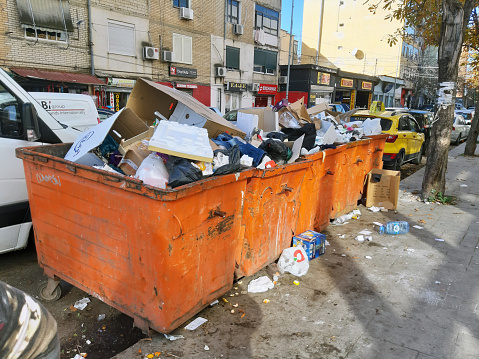 Tirana, Albania - December 20, 2023: Overflowing Garbage Container, Parking Lot, Land Vehicle, People, Retail Store, Residential Building Exterior, Taxi, Tree Scene