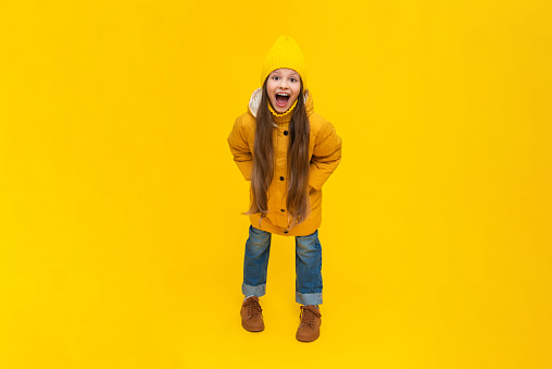 A little girl in full height in a down jacket and a hat smiles with her mouth wide open. The child is happy about the onset of autumn. Warm clothes for children. Yellow isolated background.