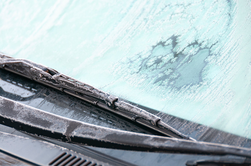 An image depicting a car's frosty windscreen with wipers, capturing the intricate patterns of ice crystals formed against the glass. This scene conveys the crispness of a cold morning, a familiar winter occurrence highlighting the season's chill.