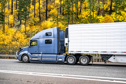 Long carrier blue big rig semi truck with extended cab for truck driver rest transporting cargo in reefer semi trailer driving on highway road with autumn forest on hills in Columbia Gorge area