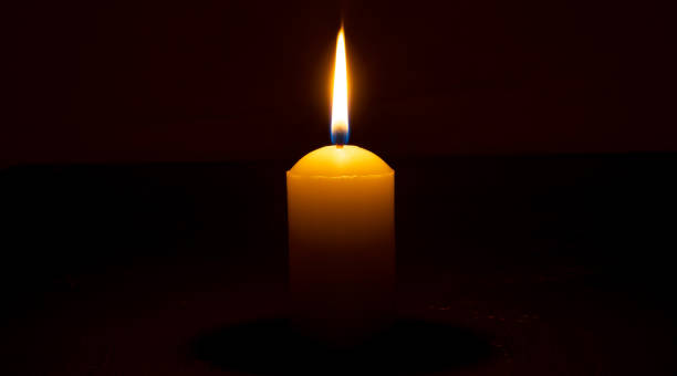 single burning candle flame or light glowing on a big white candle on black or dark background on table in church for christmas, funeral or memorial service with copy space. - candle memorial vigil praying candlelight photos et images de collection