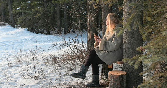 Mature woman rests on bench in forest and uses mobile phone on winter day