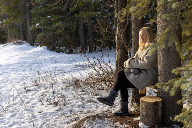 Mature woman rests on bench in forest and uses mobile phone on winter day