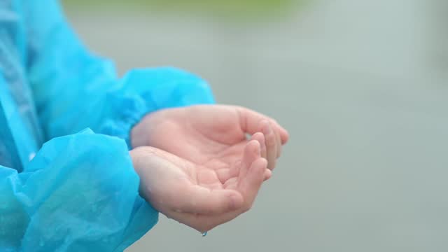 Close-up of a child's hands in the pouring rain. A joyful boy in a blue raincoat tries to catch raindrops in his palms while walking on a rainy day.