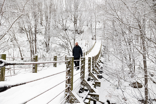 A hiker enjoying winter snowshoeing crossing a snow covered suspension bridge in a white forest.