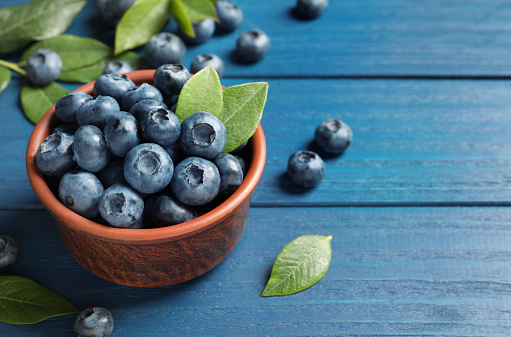 Tasty fresh blueberries in bowl on blue wooden table, space for text