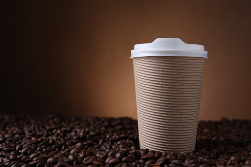 Coffee to go. Paper cup on roasted beans against brown background, space for text