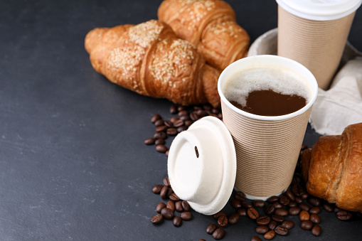 Coffee to go. Paper cup with tasty drink, croissants and beans on black table, space for text