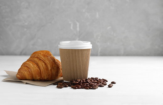 Coffee to go. Paper cup with tasty drink, croissant and beans on white wooden table. Space for text