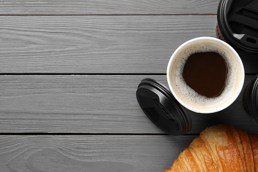 Coffee to go. Paper cups with tasty drink and croissant on grey wooden table, flat lay. Space for text