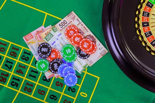An casino roulette table with Mexican peso banknotes, poker chips is displayed on green gaming surface