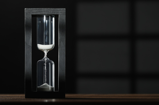Hourglass with flowing sand on wooden table against black background, space for text