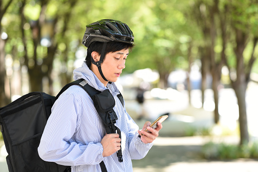 A young Asian man carrying a backpack provides a delivery service on his bicycle and searches for a delivery address on his smartphone.