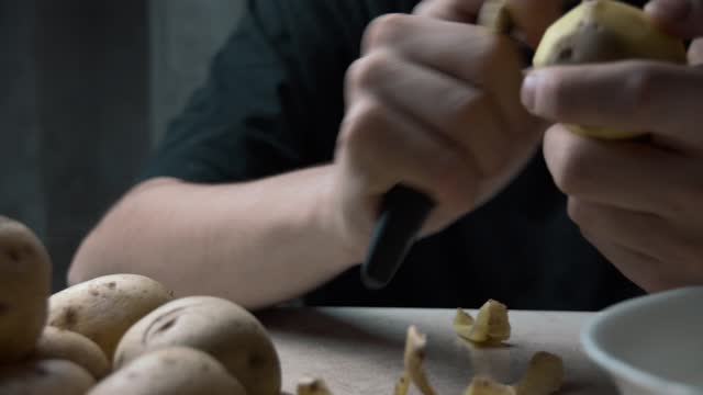 Men's hands peel potatoes at the kitchen table. A cinematic picture. The passage is from right to left.