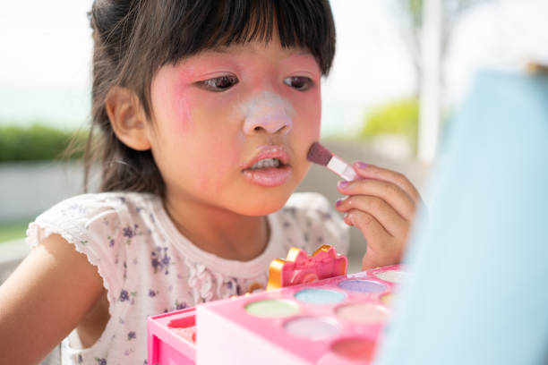 adorable little child asian girl paints her mouth with pink children heads and looks in the mirror. a child plays at home in a toy beauty salon. increase learning development for preschoolers. - child making a face human face humor - fotografias e filmes do acervo