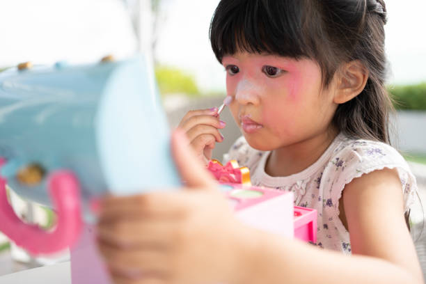 adorable little child asian girl paints her mouth with pink children heads and looks in the mirror. a child plays at home in a toy beauty salon. increase learning development for preschoolers. - child making a face human face humor - fotografias e filmes do acervo