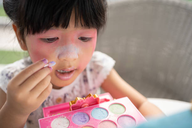 adorable little child asian girl paints her mouth with pink children heads and looks in the mirror. a child plays at home in a toy beauty salon. increase learning development for preschoolers. - smiling humor child making a face - fotografias e filmes do acervo