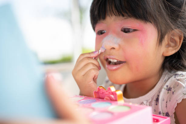 adorable little child asian girl paints her mouth with pink children heads and looks in the mirror. a child plays at home in a toy beauty salon. increase learning development for preschoolers. - smiling humor child making a face - fotografias e filmes do acervo