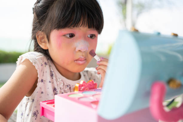 adorable little child asian girl paints her mouth with pink children heads and looks in the mirror. a child plays at home in a toy beauty salon. increase learning development for preschoolers. - child making a face human face humor стоковые фото и изображения