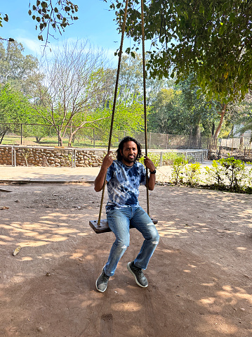 Stock photo showing a woodland swing tied to the branch of a tree by rope with Indian man swinging in dappled sunlight.