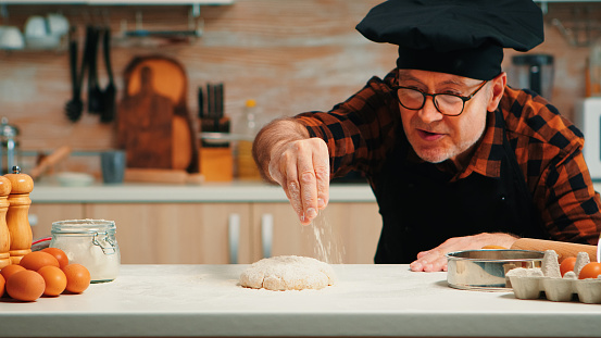 Elderly chef with bonete making dough baking cookies. Retired senior baker with apron, kitchen uniform sprinkling, sifting, spreading rew ingredients with hand baking homemade pizza and bread.