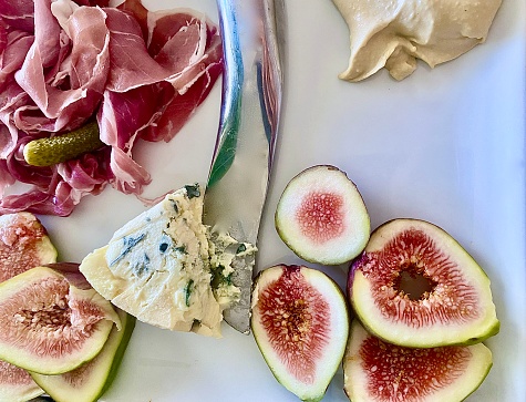 Horizontal close up flat lay of white appetiser potter with prosciutto, blue vein cheese, fresh figs, humous, and silver cutting knife in domestic kitchen